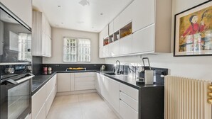A modern kitchen with white cabinets, black countertops, and a built-in oven. There is a window with blinds, a radiator, and a framed artwork on the wall.