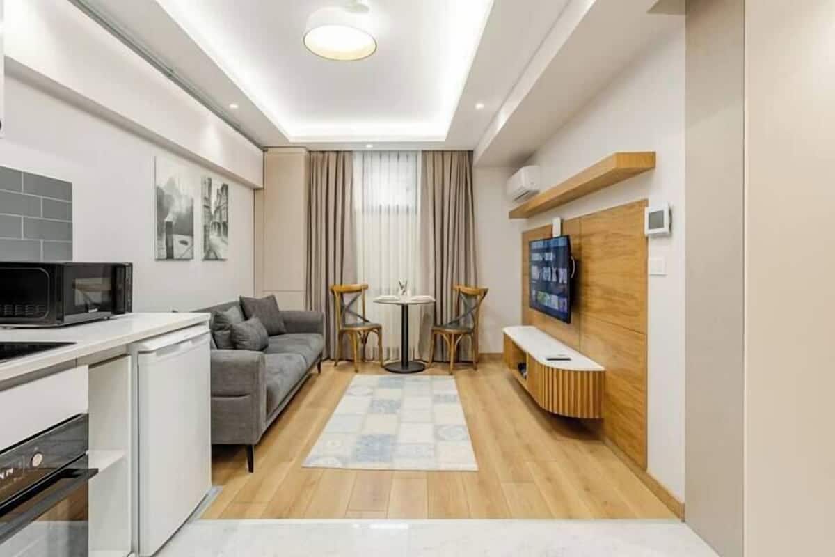 A compact living space with a kitchenette, a grey sofa, a small round table, and a TV mounted on the wall.