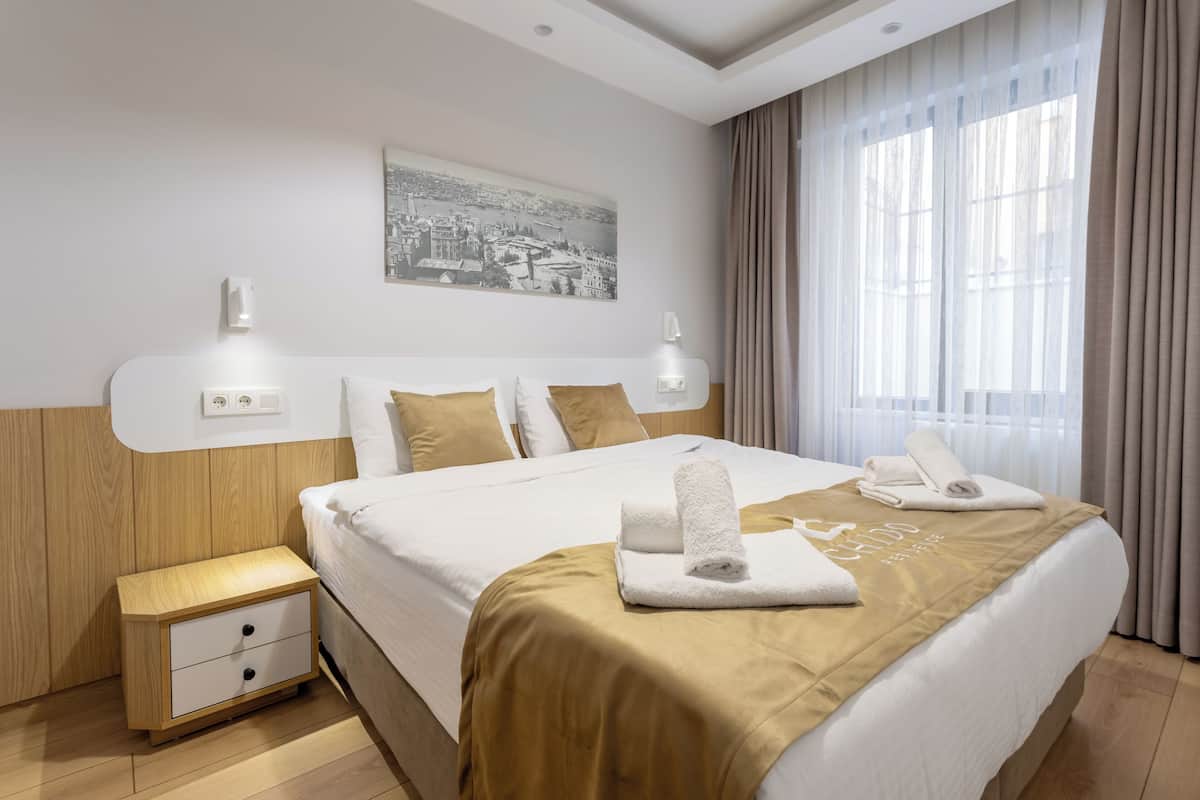 A hotel room with a neatly made bed, a nightstand with a drawer, a wall-mounted light, and a window with curtains.