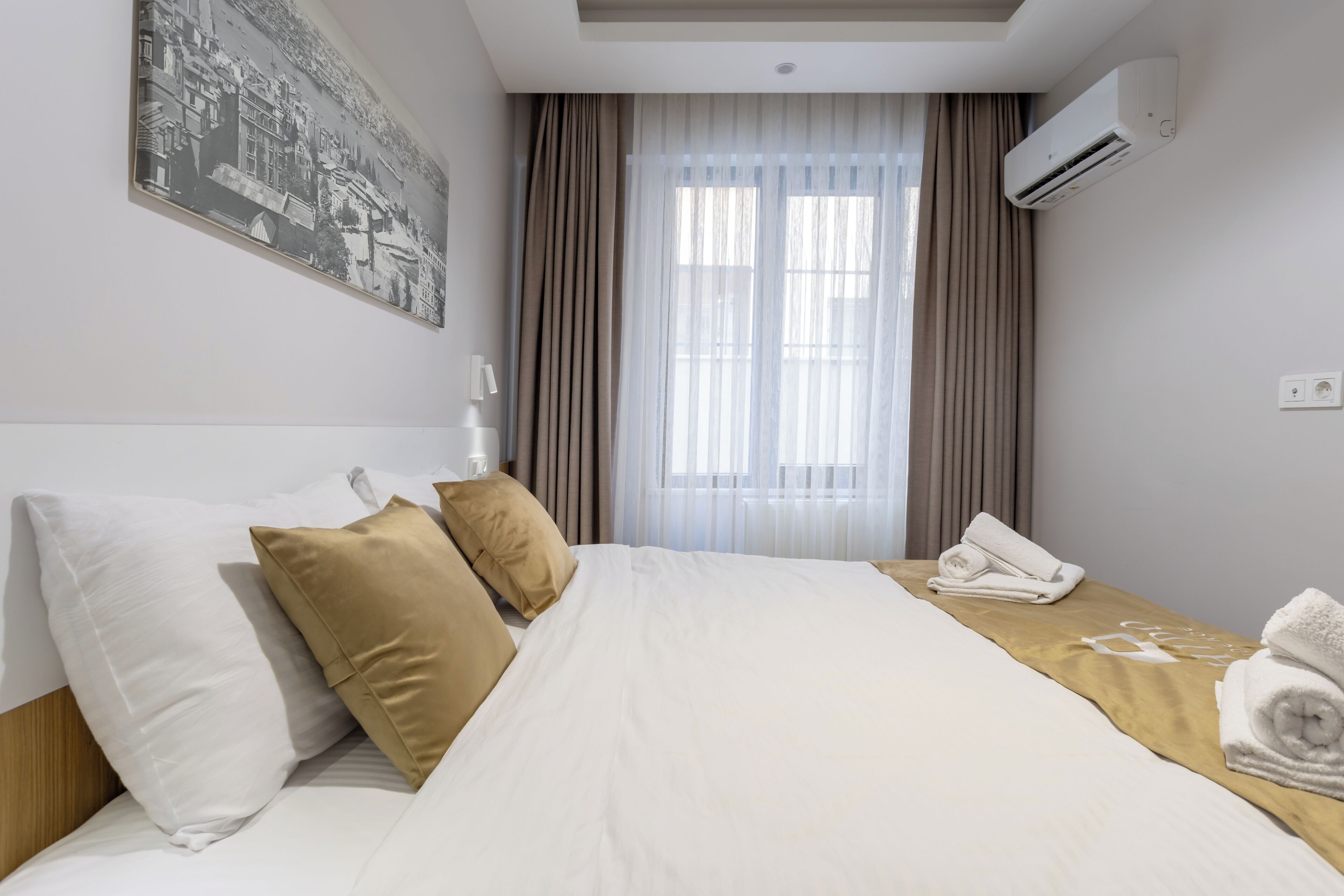 Une chambre d’hôtel avec un grand lit, des oreillers beiges et blancs, un lit bien fait avec une serviette, un appareil de climatisation et une fenêtre avec des rideaux.