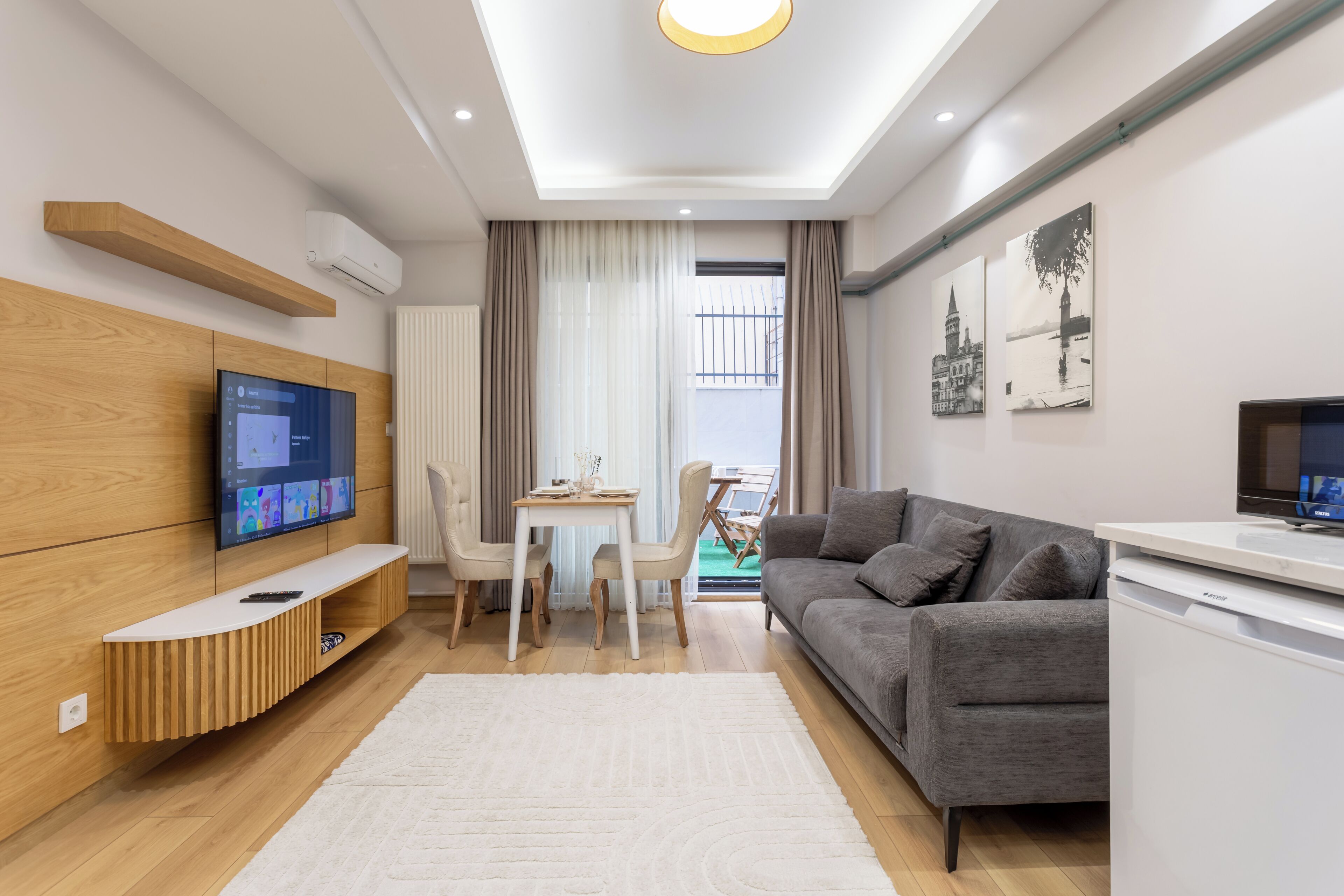 A modern living room with a flat-screen TV, a grey sofa, a dining table with chairs, and a white rug.