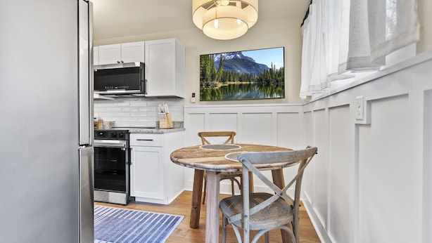 A kitchen with white cabinets, a wooden dining table with chairs, a large window with curtains, and a painting of a mountain lake. - Abode 81 in Newark (Newark)