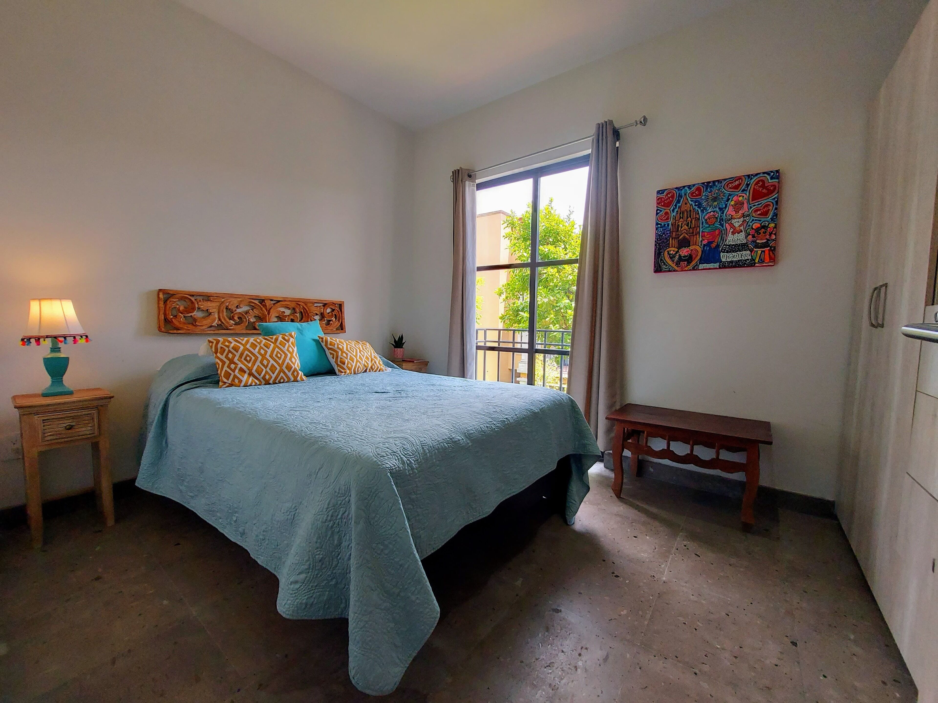 Ein Schlafzimmer mit einem Bett, einem Nachttisch, einer Bank, einem Fenster mit Vorhängen und einer Wand mit einem farbenfrohen Gemälde.