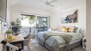 Quarto com cama, escrivaninha com computador, cadeira e varanda com vista para árvores e edifícios.