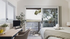 A hotel room with a balcony, a desk with a TV, a bed with a blanket, and a potted plant.