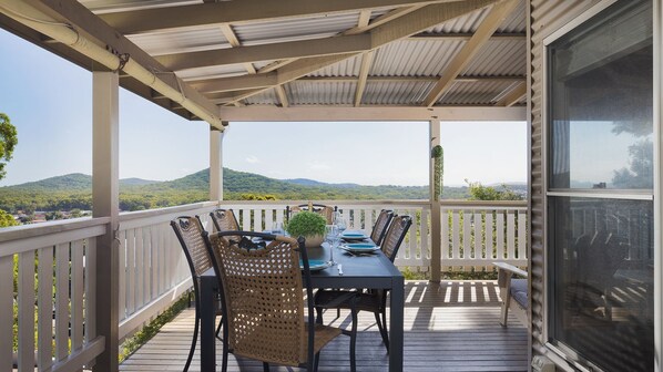 Uma área de refeições ao ar livre coberta, com uma mesa posta para quatro pessoas, cercada por cadeiras de vime, com vista para uma paisagem montanhosa.