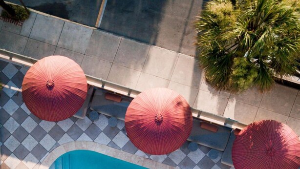 Eine Vogelperspektive auf einen Poolbereich mit gefliestem Boden, umgeben von mehreren roten Sonnenschirmen, und einem Pool mit blauer Innenseite.