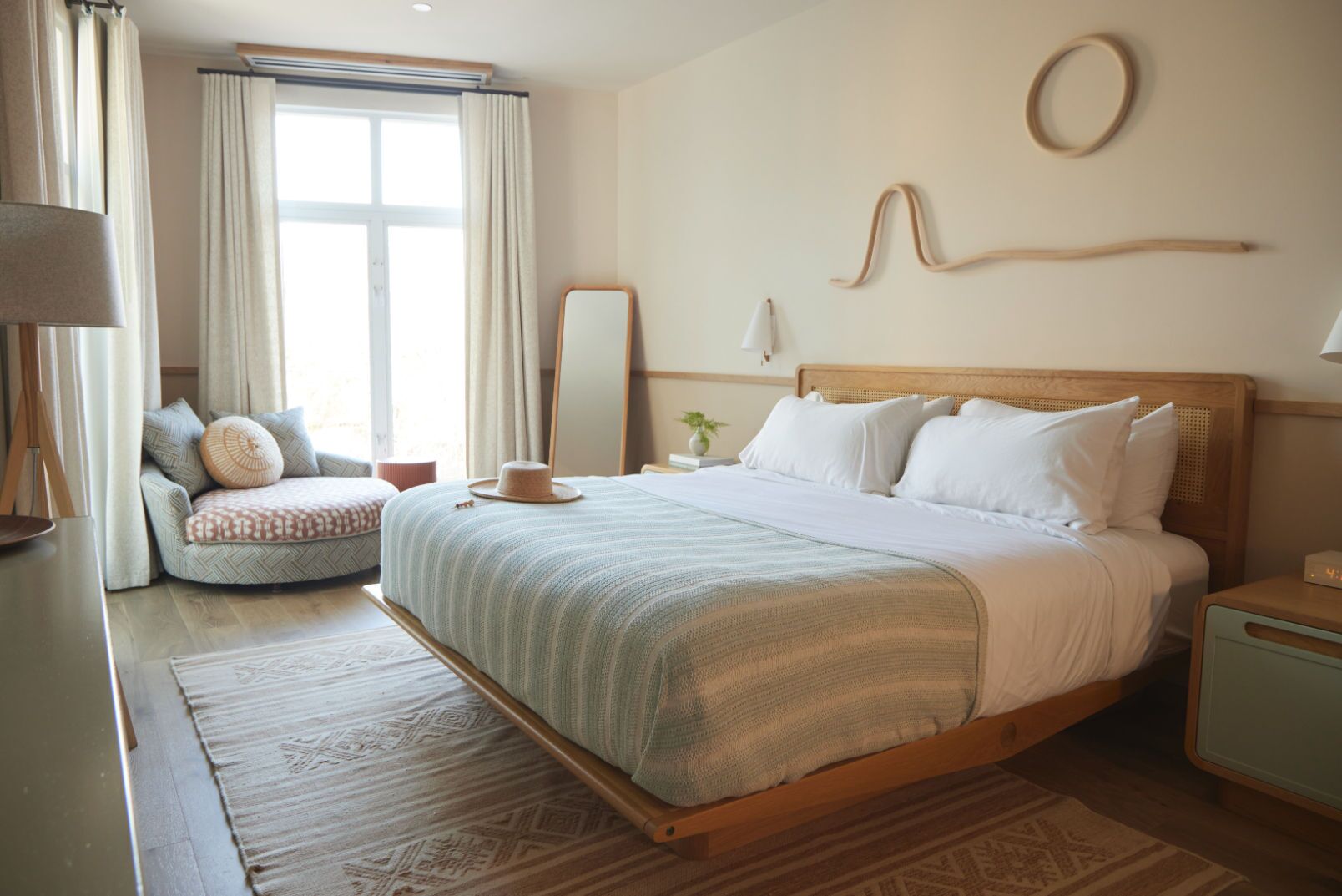 Une chambre à coucher avec un grand lit, une table de chevet, une commode, une chaise, un miroir et une fenêtre avec des rideaux.