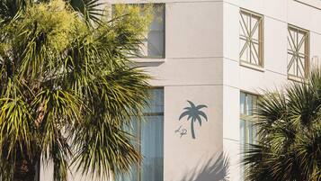 Un bâtiment avec un palmier au premier plan, un parasol rouge et une pancarte sur laquelle on peut lire : « SURVEILLANCE : BAIGNEZ-VOUS À VOS PROPRES RISQUES ».