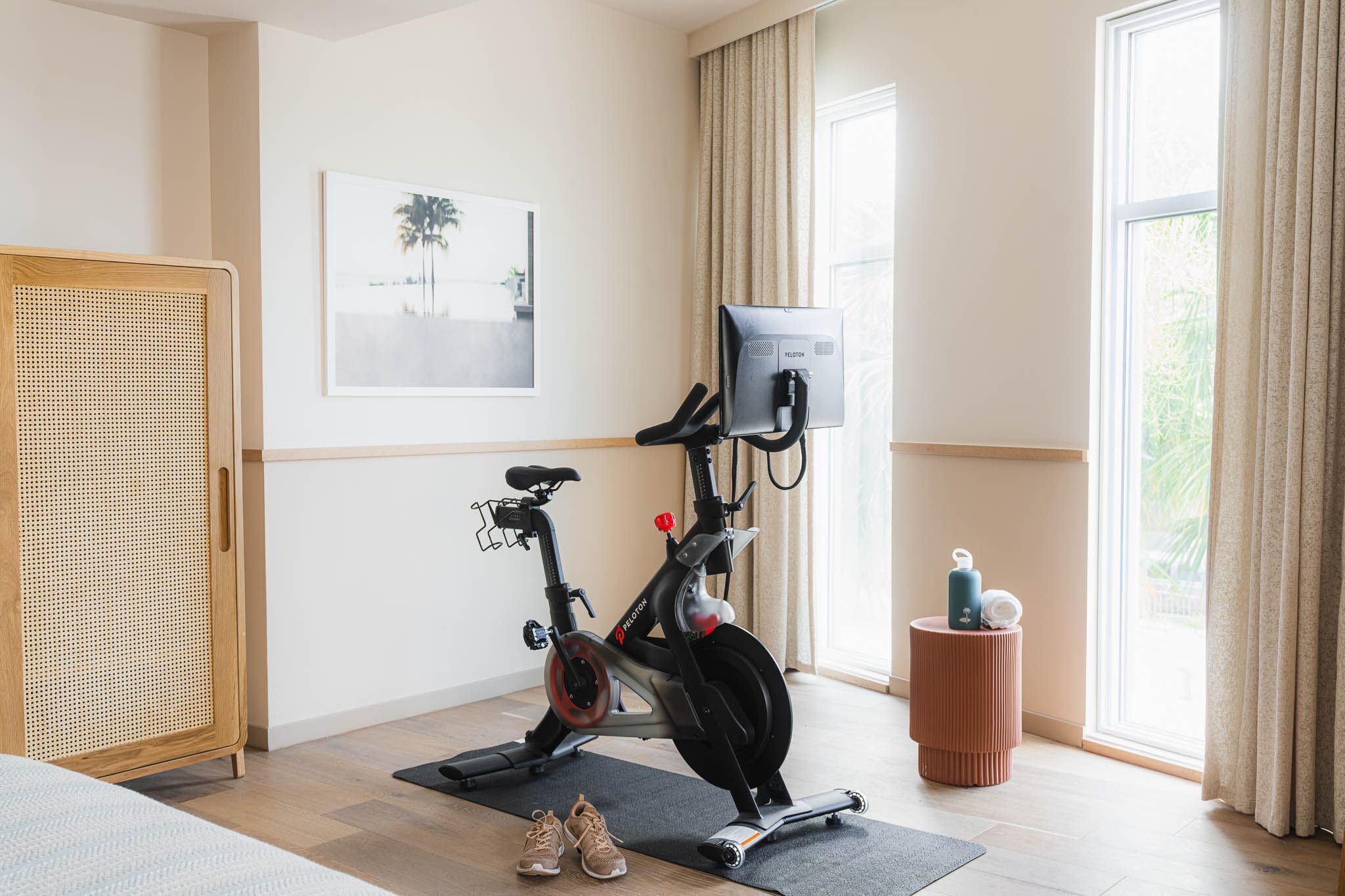 Una habitación con una bicicleta estática, un armario de ratán, un taburete pequeño y una ventana con cortinas.