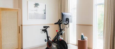 Una habitación con una bicicleta estática, un armario de ratán, un taburete pequeño y una ventana con cortinas.