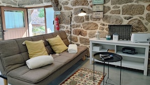 A cozy living room with a stone wall, a sofa with cushions, a small table with a lamp, and a shelf with various items. - Aldeia de Pontes (Melgaco)