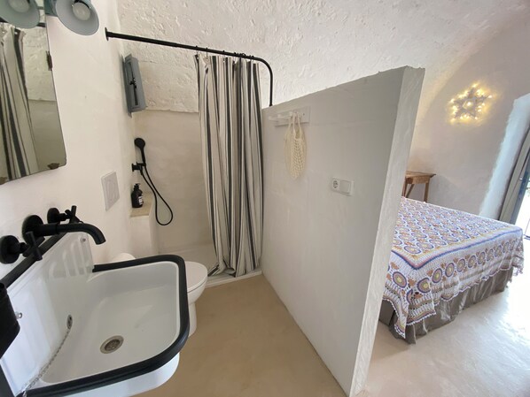 A bathroom with a sink, toilet, and shower. There is a bed with a crocheted blanket in the adjacent room.