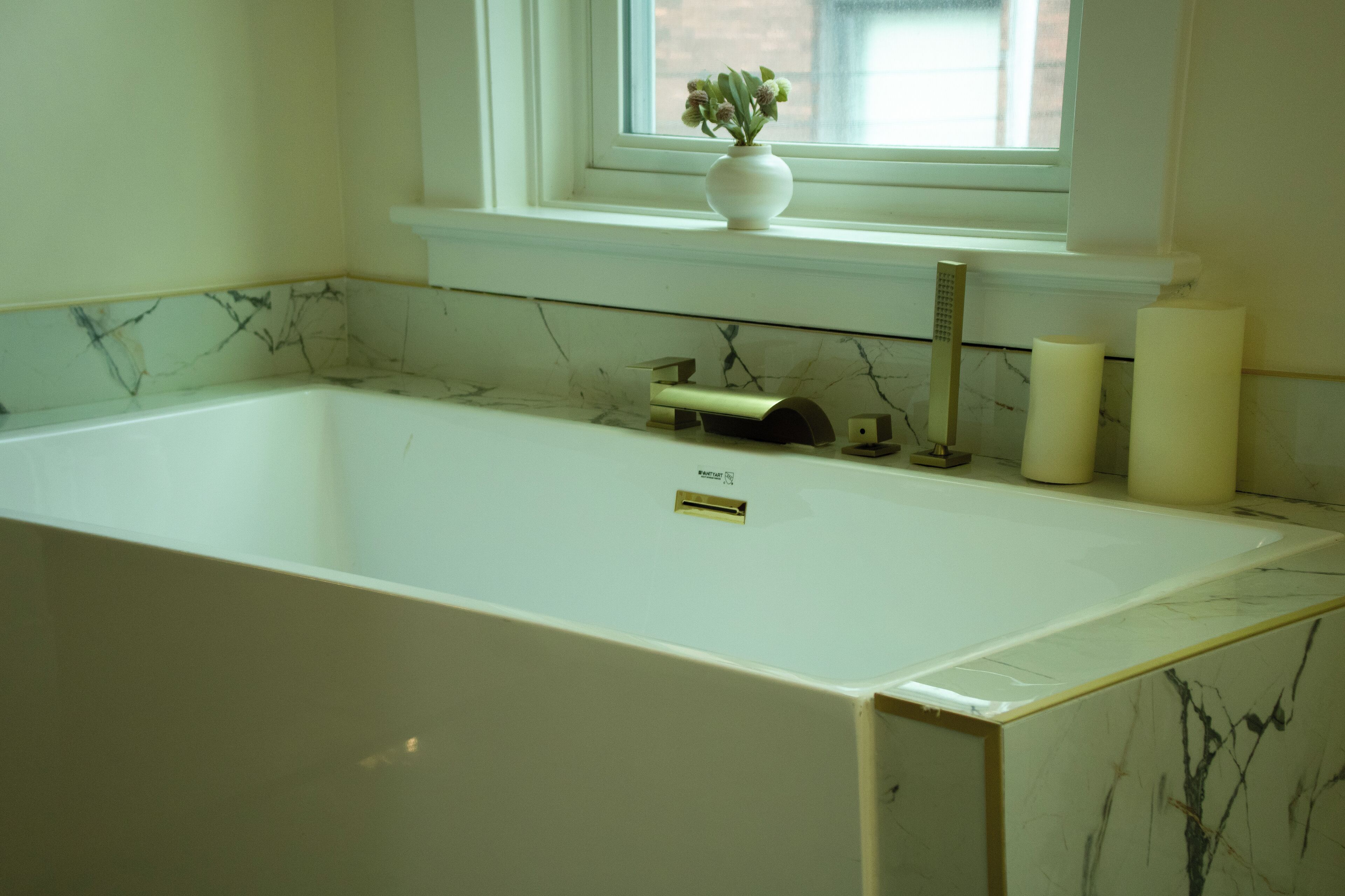 Une salle de bain avec une baignoire blanche, un pare-baignoire en marbre, une fenêtre avec une plante et deux bougies posées sur le comptoir.