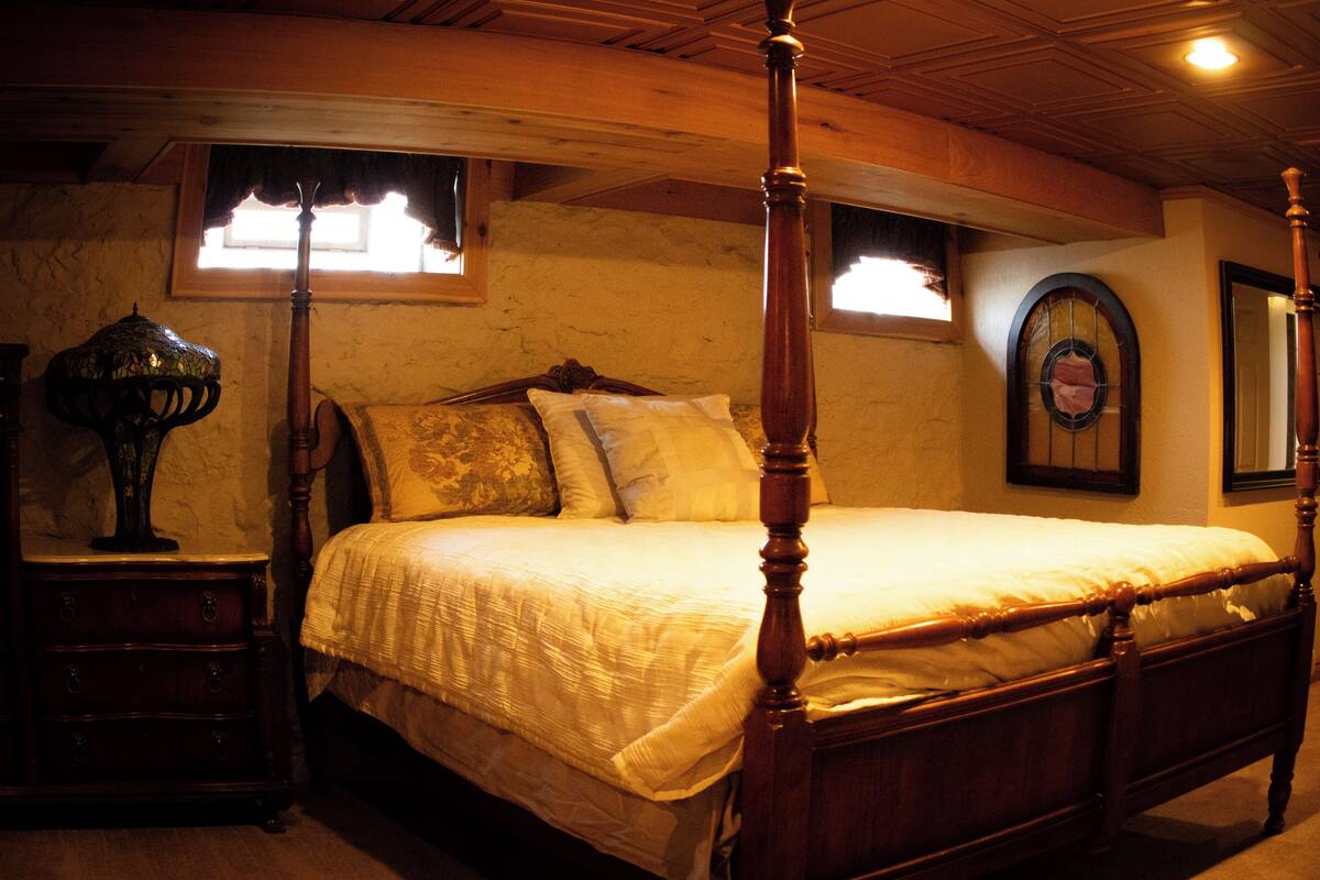 Un lit à baldaquin avec une structure en bois et un couvre-lit blanc, une table de chevet avec une lampe, et un mur avec une fenêtre et un miroir.