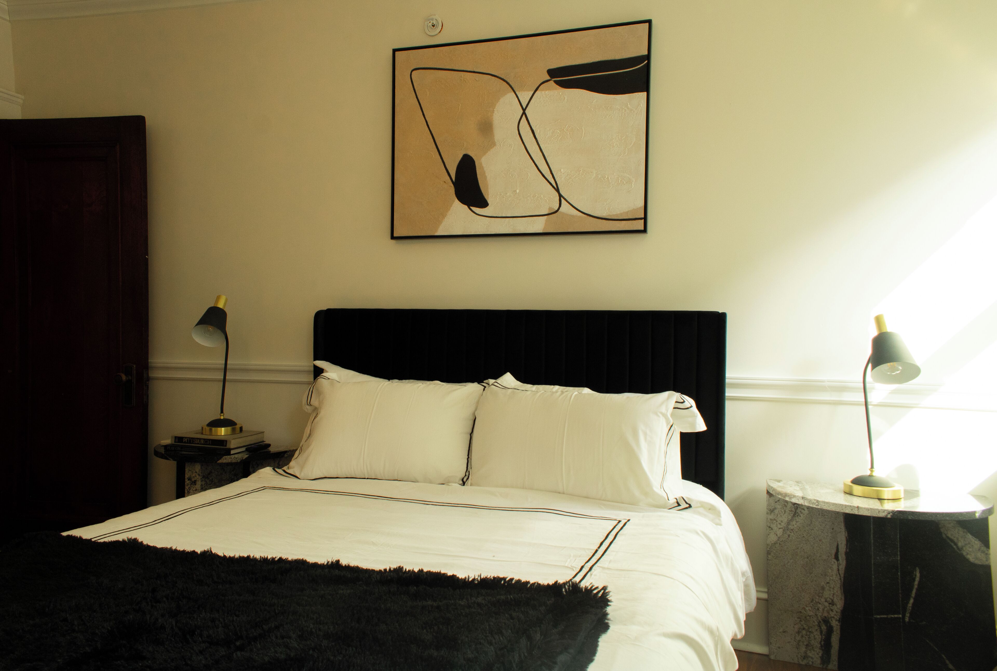 A bedroom with a bed, two bedside tables, a wall-mounted abstract painting, and a window with natural light.