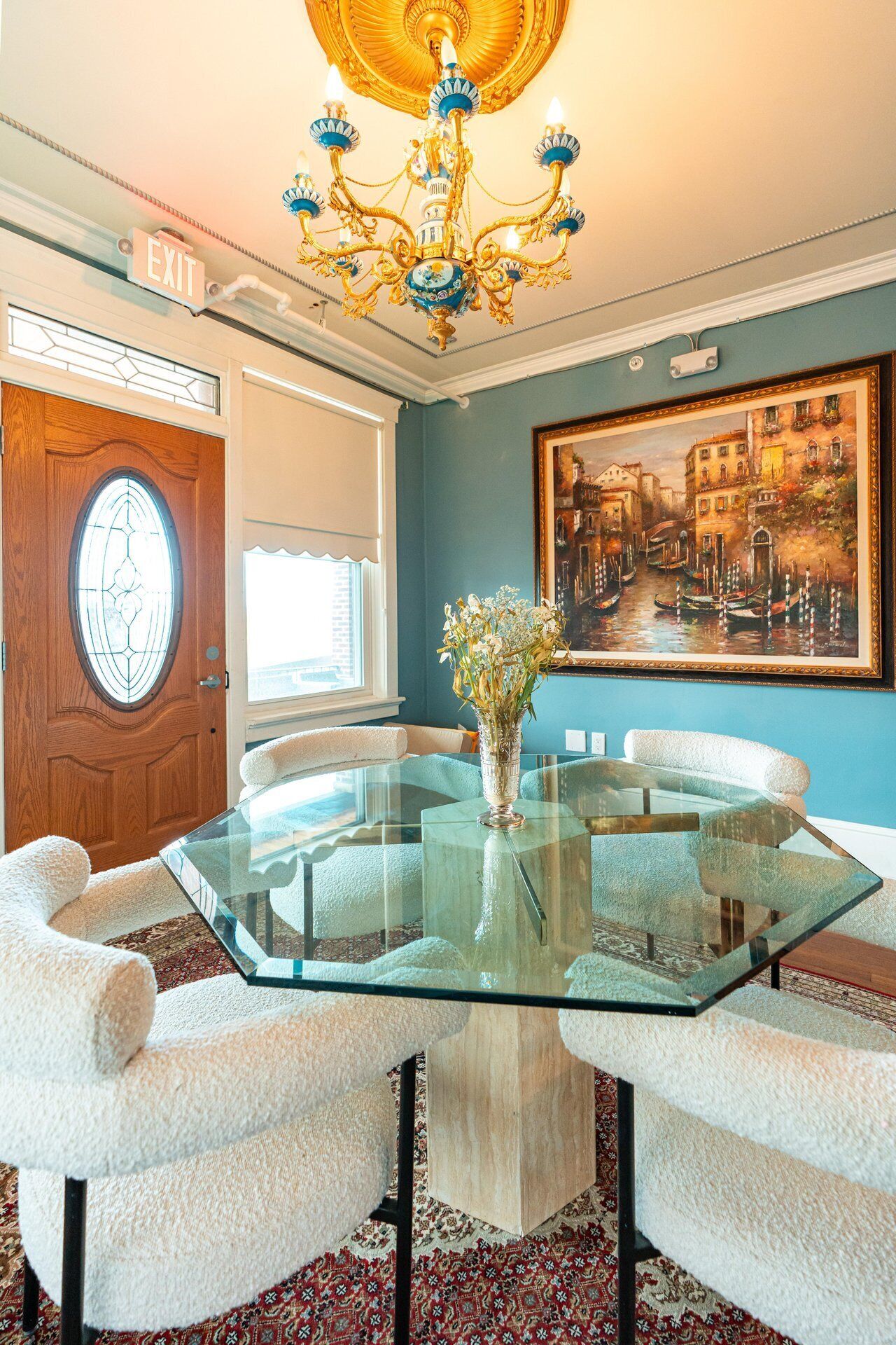 A dining area with a glass table surrounded by white chairs, a chandelier, a painting of a cityscape, and a door with a stained glass window.