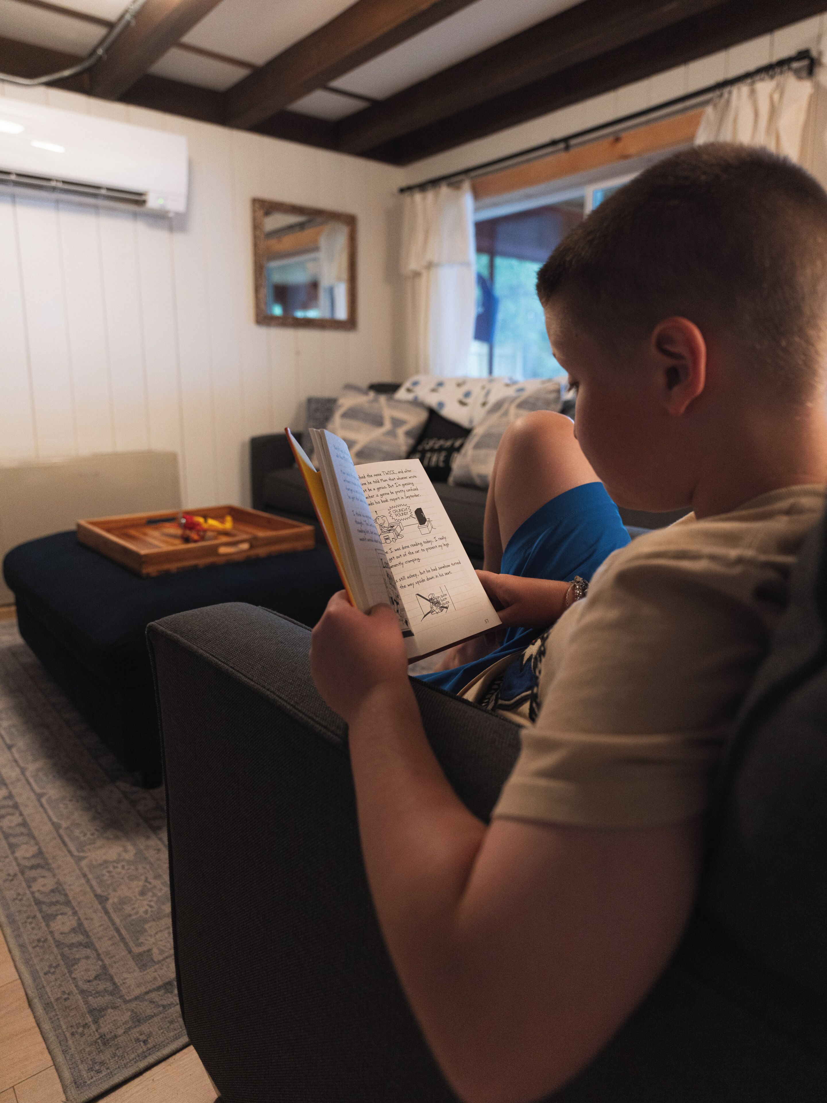Una persona sentada en un sofá leyendo un libro en una habitación con techo de madera, una ventana con cortinas y una mesita con una bandeja.