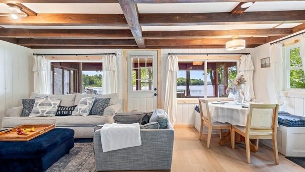 A cozy living room with a sofa, ottoman, and dining table with chairs. There are windows with curtains, a wooden ceiling, and a view of the outdoors. - Salt water hot tub, king size beds, dog friendly, lake front, dock, boat launch (Canaan)