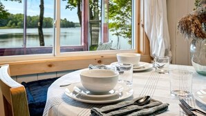 A dining table set with white plates, bowls, and cutlery, with a view of a lake and trees through large windows. - Salt water hot tub, king size beds, dog friendly, lake front, dock, boat launch (Canaan)