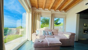 Ein modernes Wohnzimmer mit großem Fenster, einem beigen Sofa und Blick auf Grünflächen und eine Steinmauer.