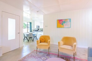 A modern living room with two yellow armchairs, a colorful abstract painting, and a dining area with a table and chairs.