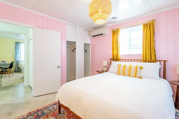 A bedroom with a bed, a bedside table, a lamp, a window with curtains, and an air conditioning unit.