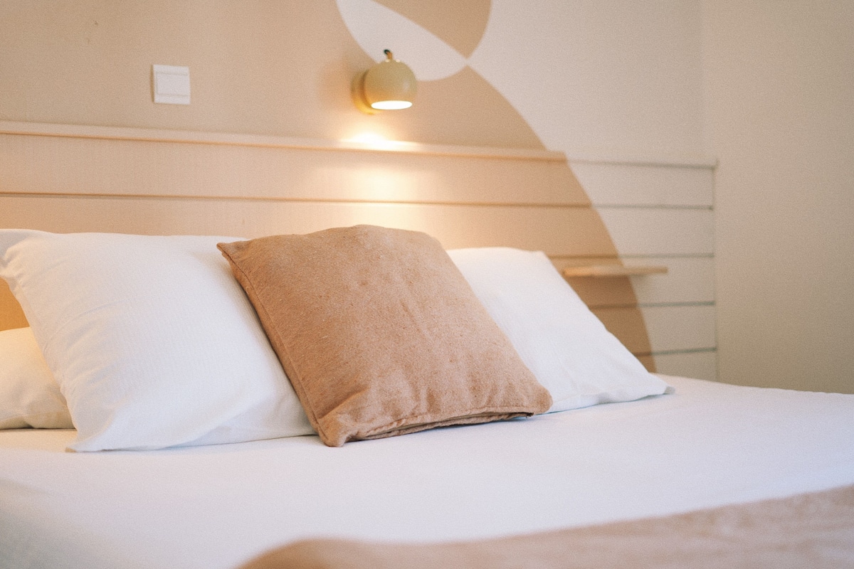 A neatly made bed with white linens and a brown towel, a wall-mounted light fixture, and a simple, clean room design.
