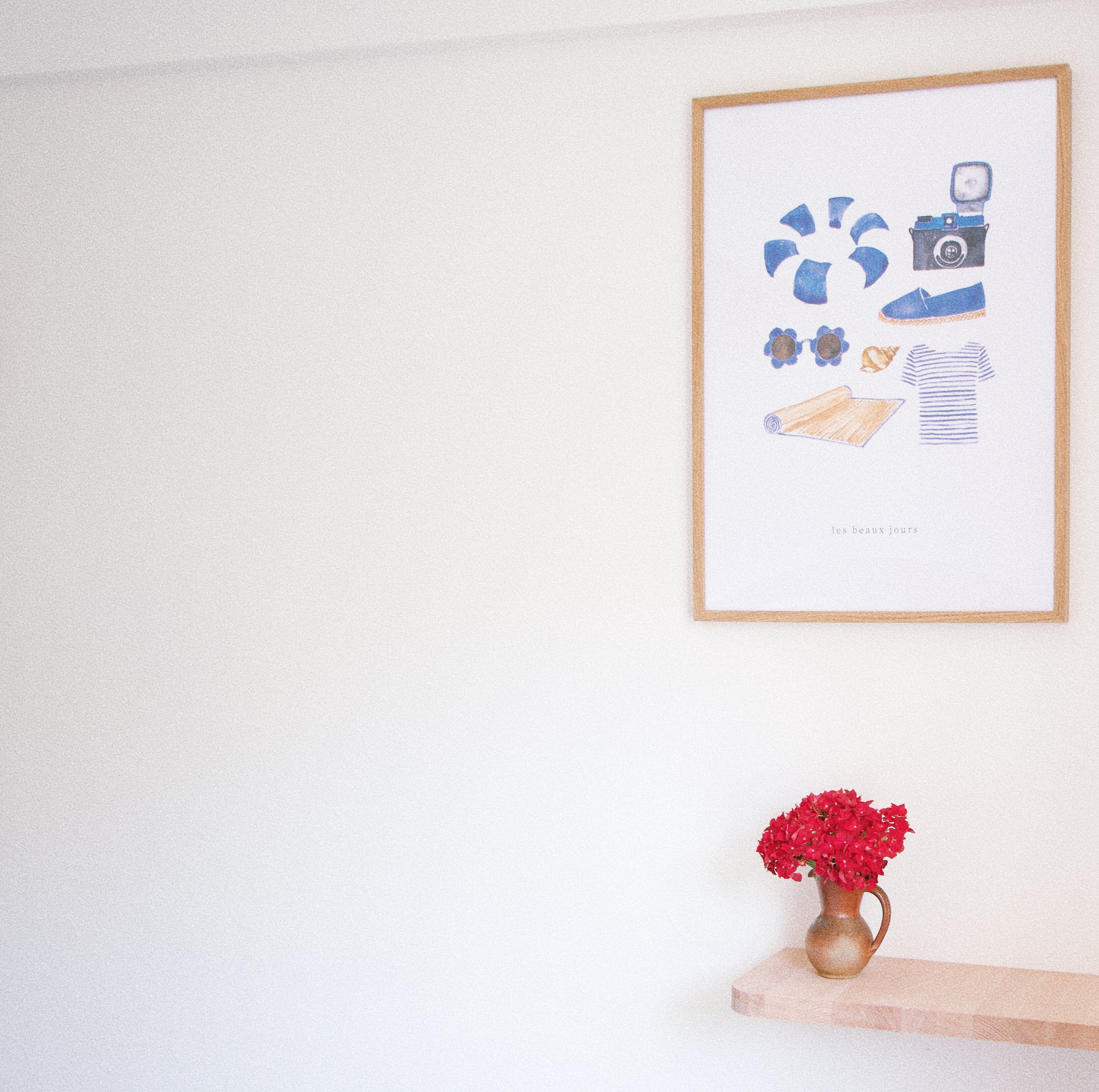 Une œuvre d’art encadrée accrochée au mur, un vase avec des fleurs rouges posé sur une étagère en dessous, et un plancher en bois.