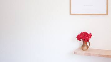 Une œuvre d’art encadrée accrochée au mur, un vase avec des fleurs rouges posé sur une étagère en dessous, et un plancher en bois.