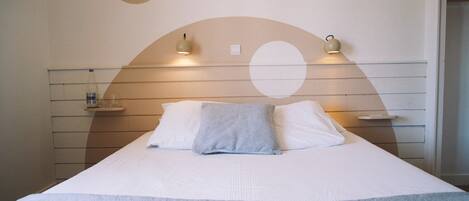 Ein ordentlich gemachtes Bett mit weißer Decke und blauer Tagesdecke, zwei Nachttischlampen und eine Wand mit geometrischem Muster.