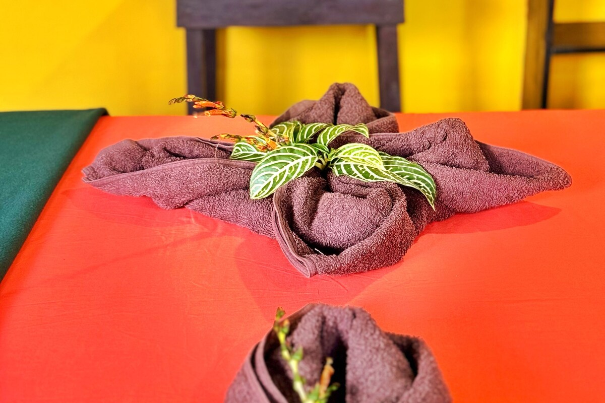A neatly made bed with a red bedspread and a brown towel with a plant.