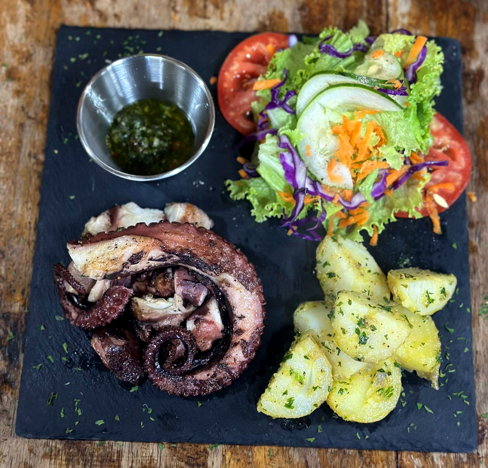 A meal consisting of grilled octopus, a salad with tomatoes, cucumbers, and purple cabbage, and roasted potatoes, served on a slate board.