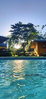 Piscina com águas cristalinas refletindo o sol, rodeada por vegetação e um edifício com telhado em azulejos.