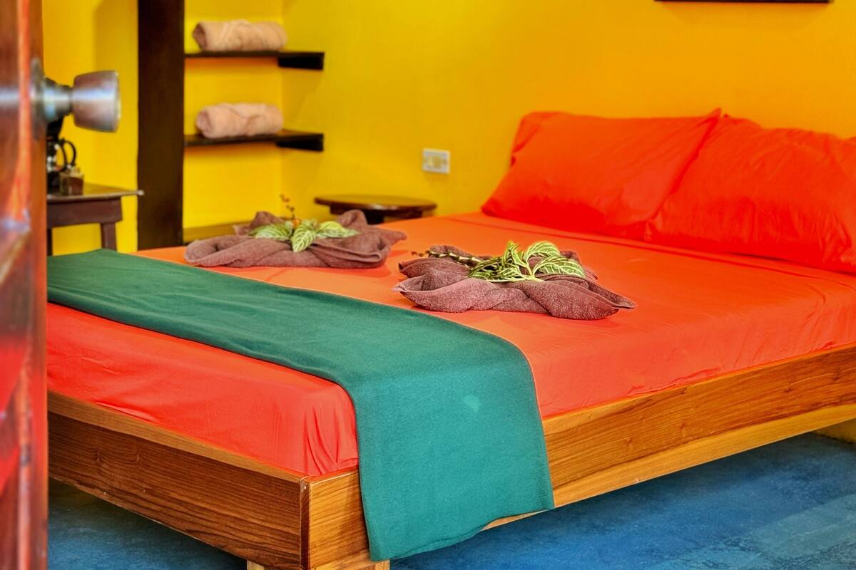A room with a bed, a nightstand, a shelf with towels, and a painting on the wall.