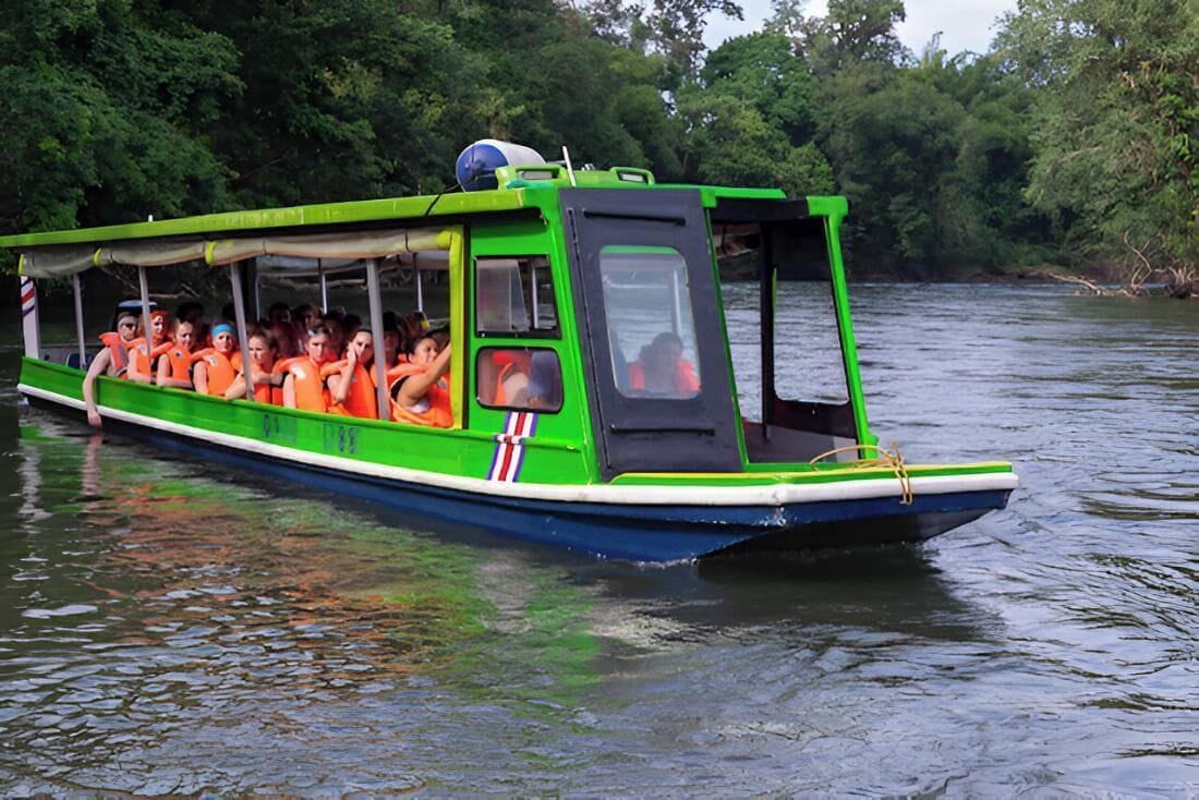 Una embarcación verde y azul con pasajeros que llevan chalecos salvavidas anaranjados, navegando por un río rodeado de vegetación.