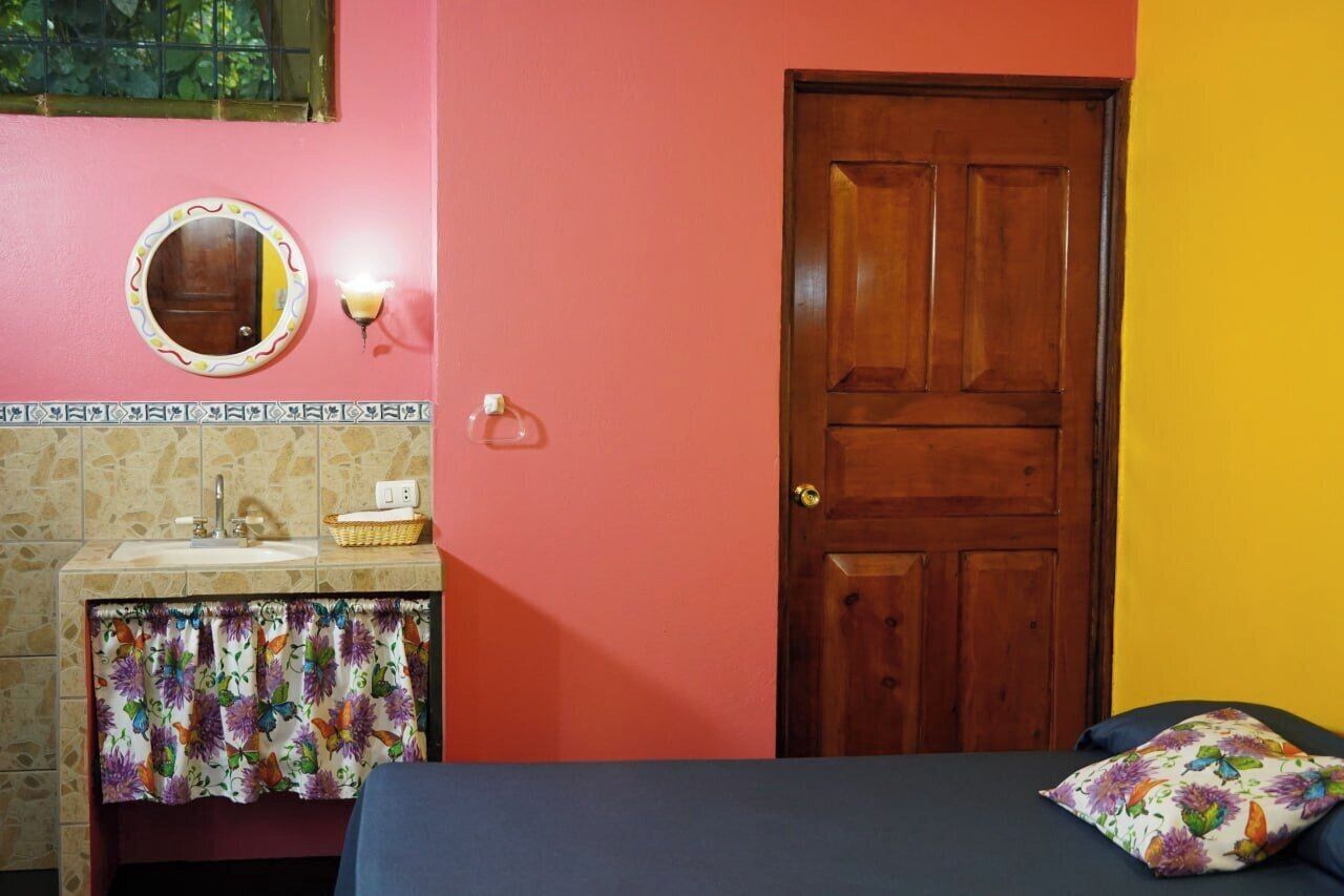 A room with a wooden door, a mirror, a sink with a floral curtain, and a bed with a floral pillow.