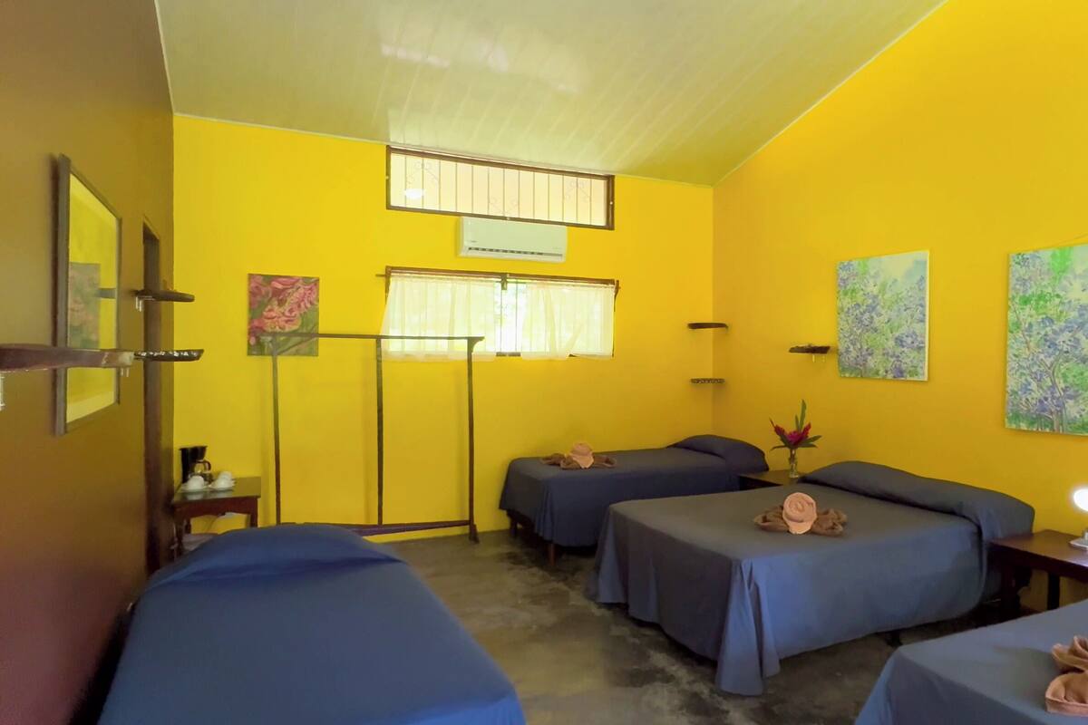 A room with four beds, a window with curtains, a small table with a lamp, and two paintings on the wall.