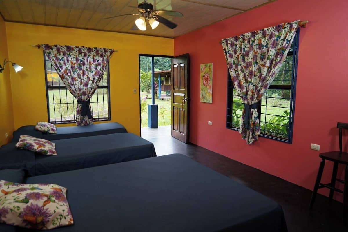 A room with three beds, floral curtains, a ceiling fan, and a view of the outdoors.