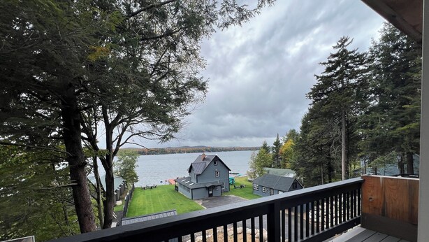 Una terraza de madera con barandilla que da a un lago, rodeada de árboles y una casa con muelle.