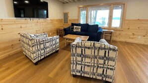 Una habitación acogedora con paredes de madera, un televisor de pantalla plana grande, dos sofás estampados, una mesita de café y una ventana con vistas a árboles.