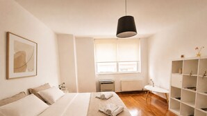 Une chambre à coucher au style minimaliste, avec un lit, un radiateur, une fenêtre et une étagère blanche.