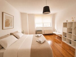 Un dormitorio minimalista con cama, radiador, ventana y una estantería blanca.