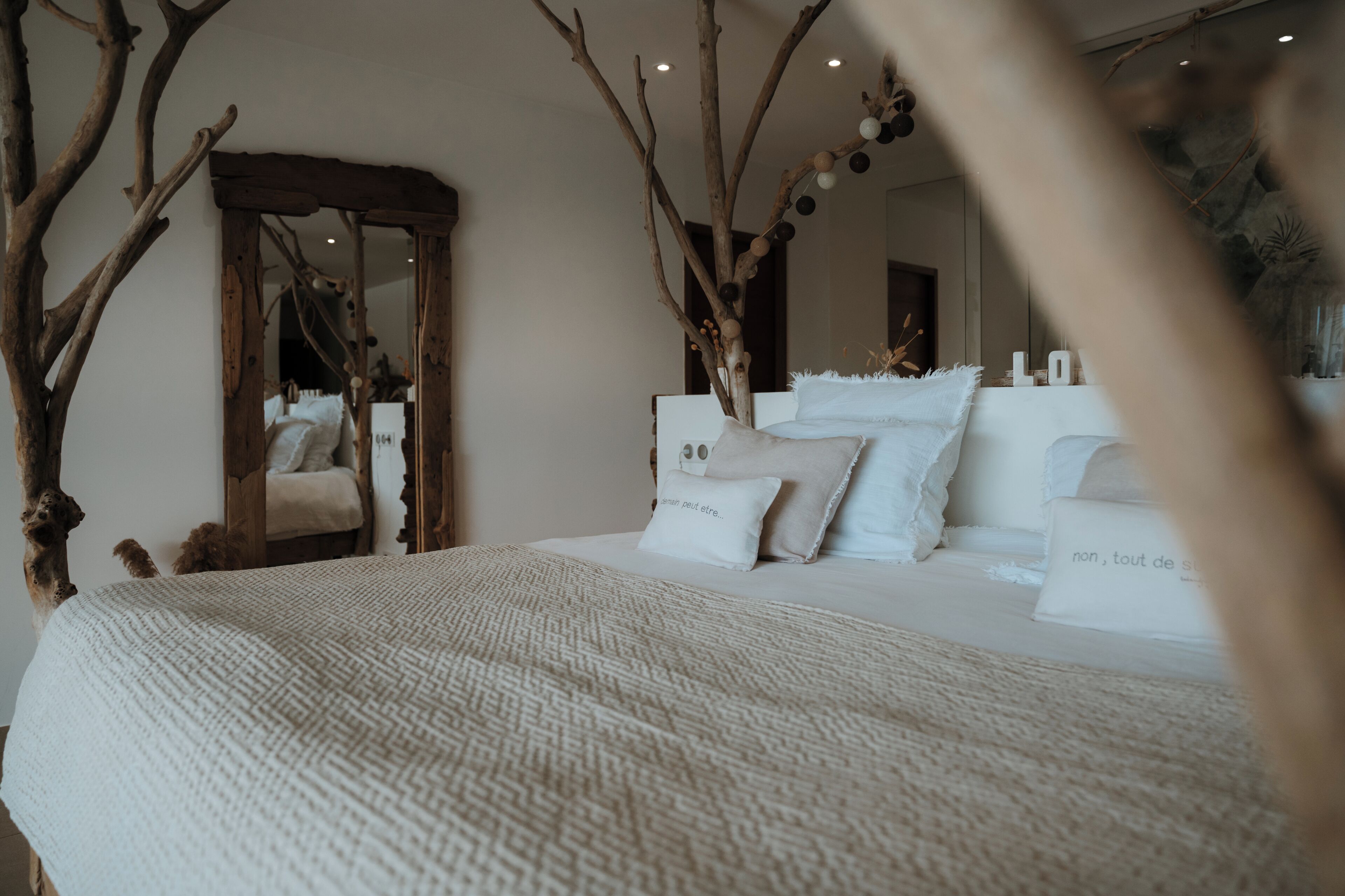 A bedroom with a large bed, a wooden tree branch decor, and a mirror reflecting the room.