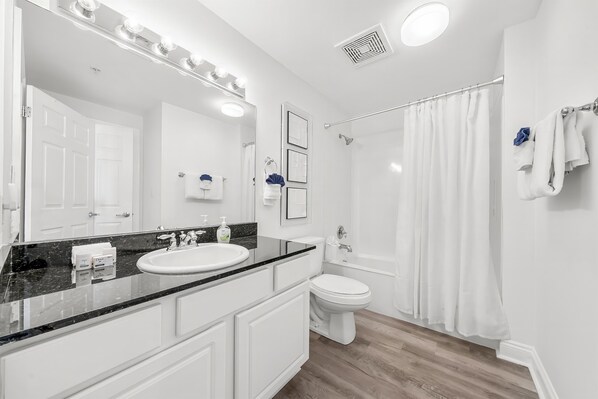 A modern bathroom with a white sink, vanity, and toilet. It includes a shower with a curtain, a large mirror with lights, and a door with a towel rack.