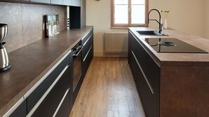 A modern kitchen with dark cabinets, a wooden floor, a built-in island with a sink, and a window with blinds. - Apartment Schöne Schwarzwald Auszeit in the hiking paradise with its own terrace (Friedenweiler)