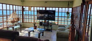 Un salon avec une grande télévision, une table basse en verre et un canapé. De grandes fenêtres offrent une vue sur l’océan.