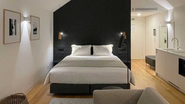 A modern bedroom with a large bed, a dark accent wall, a small kitchenette, and a bathroom visible in the background. - Studio.Ø5 City View (Porto)