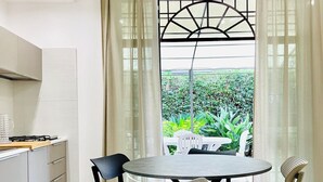 Une cuisine moderne avec une table à manger ronde et des chaises, une cuisinière à gaz et une vue sur un jardin, visible à travers une fenêtre ornée d’une grille métallique.