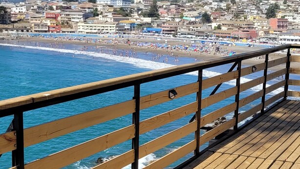 Une jetée en bois, dotée d’une rambarde, surplombe une plage où les vagues déferlent.